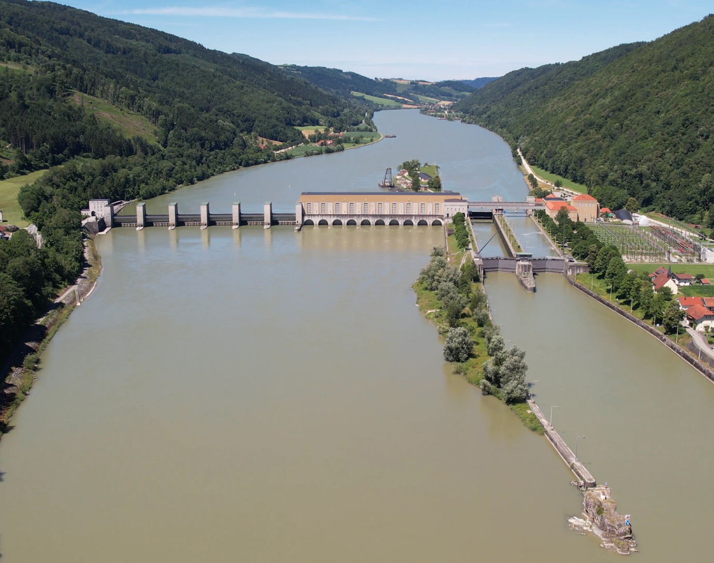 Zu sehen ist das Kraftwerk Jochenstein aus der Luft an einem sonnigen Tag. Während die Donau grünblau gefärbt ist, strahlen an den Ufern links und rechts die Bäume im saftigen Dunkelgrün. Während links die Wehrfelder zu sehen sind, sieht man rechtsseitig die Schleuse für die Schifffahrt.  Im Hintergrund sieht man noch die Landschaft sowie den Verlauf der Donau flussaufwärts.