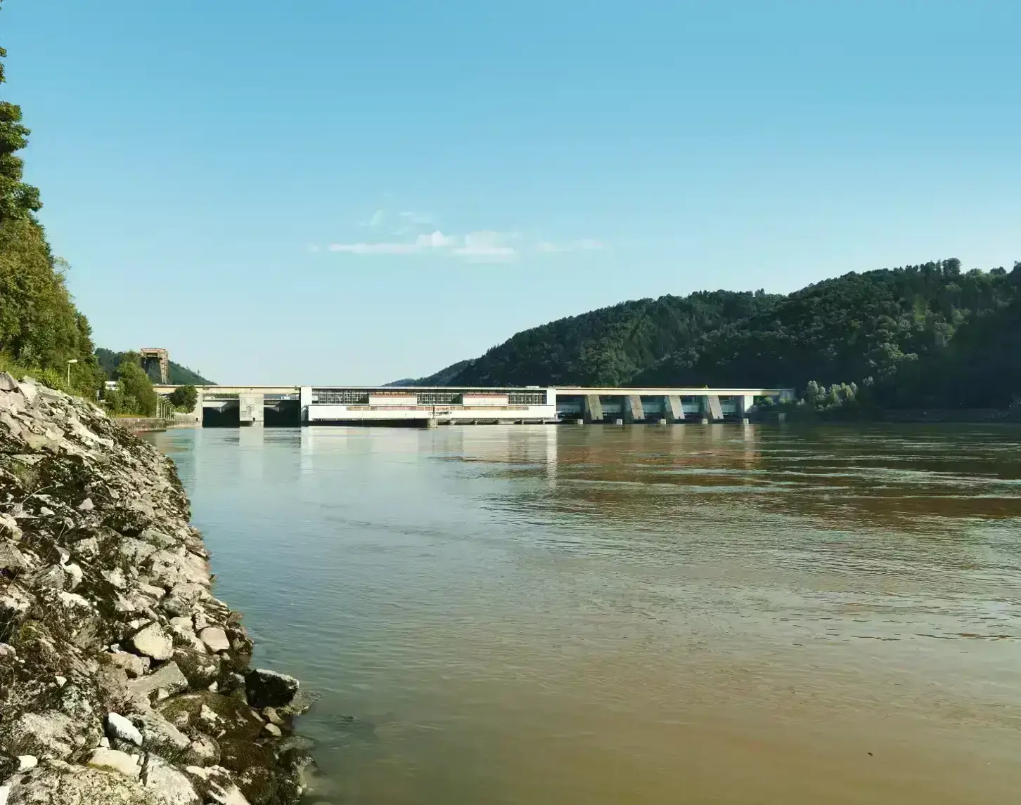 The Aschach power station can be seen from the downstream side on a beautiful summer's day. On the left you can see rocks and green trees above. In front of the power station you can see the greenish Danube flowing downstream.  To the right of the power station you can see dark green forests and behind the power station you can see a bright blue sky.