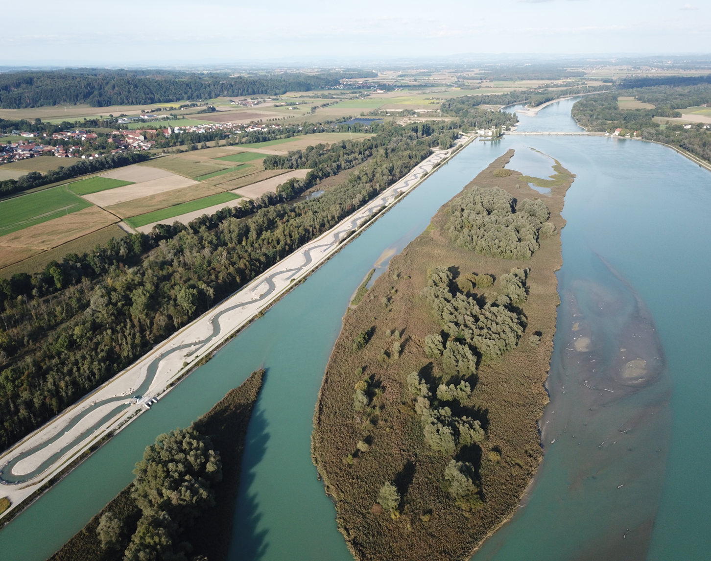 Das Luftbild zeigt den Innabschnitt vor dem Innkraftwerk Ering-Frauenstein. Im linken Bereich des Bildes ist neben einem Waldabschnitt die Fischwanderhilfe zu sehen. In der Bildmitte befindet sich der renaturierte Bereich des Inns, welcher in einem dunklen Blau erstrahlt. Der renaturierte Bereich ist voll bewachsen mit Bäume und Sträuchern. Im Hintergrund des Bildes sieht man den weiteren Verlauf des Inns sowie die Landschaft mit Ortschaften und Feldern in verschiedensten Farben.