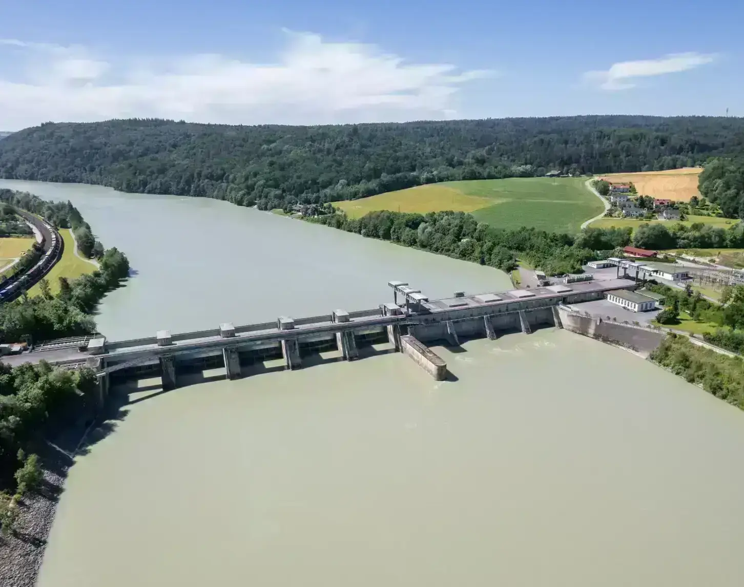 The picture shows an aerial view of the Passau-Inglings power station from the downstream side. The Inn is blue-green and therefore stands out strongly against the light and dark green meadows and forests to the left and right of the river. The course of the Inn can still be seen in the background, with forest and blue sky behind it.