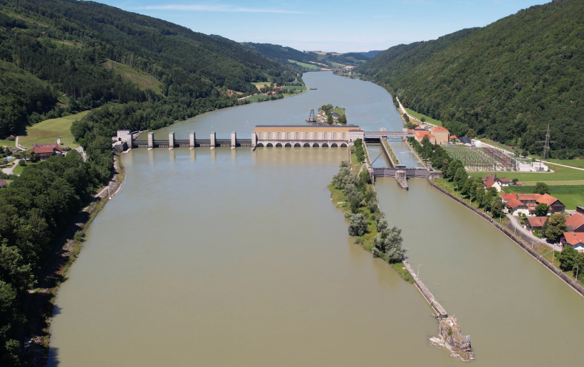 The picture shows an aerial view of the Passau-Inglings power station from the downstream side. The Inn is blue-green and therefore stands out strongly against the light and dark green meadows and forests to the left and right of the river. The course of the Inn can still be seen in the background, with forest and blue sky behind it.