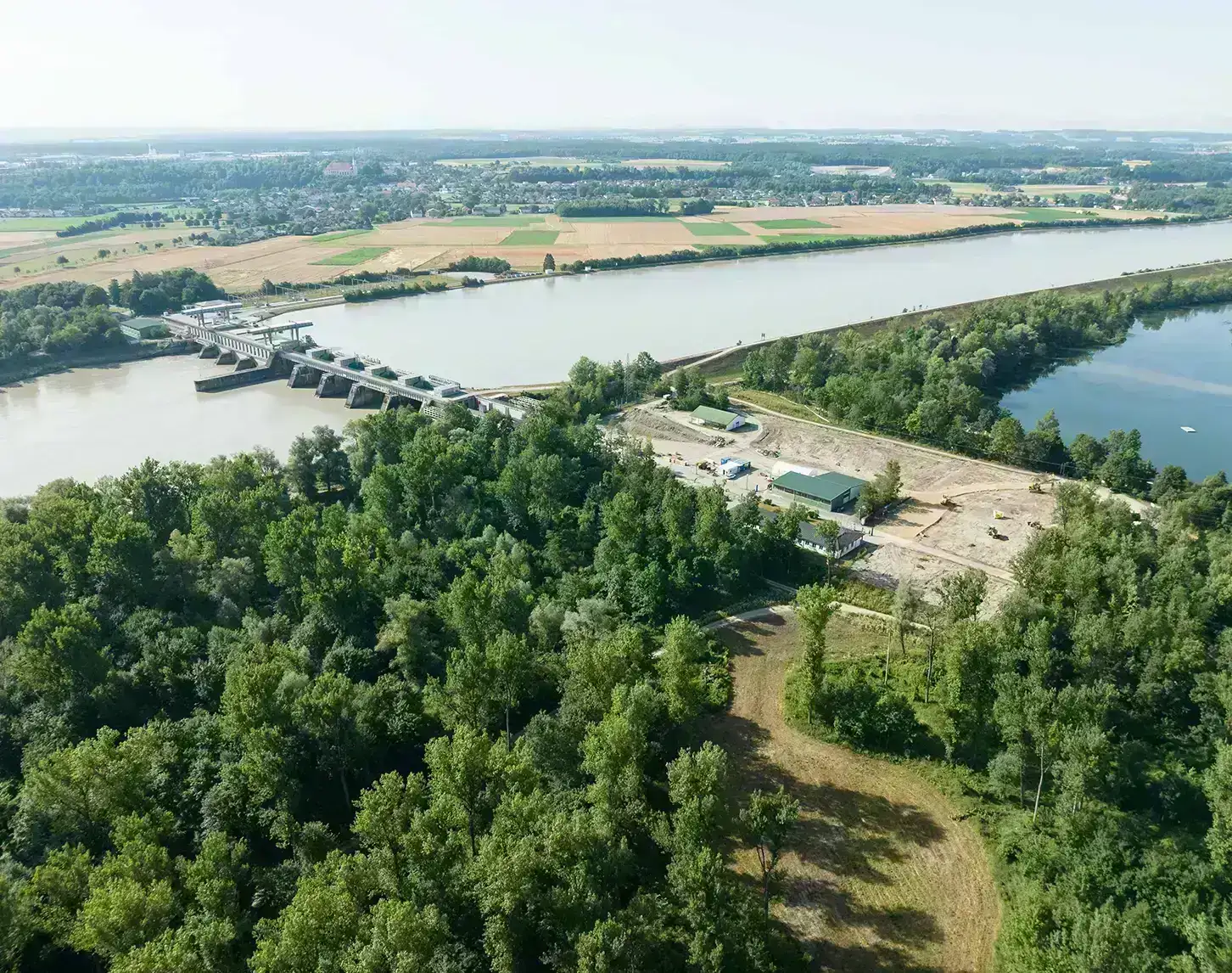 Luftaufnahme eines Wasserkraftwerks am Fluss mit einem Waldgebiet im Vordergrund und landwirtschaftlichen Feldern im Hintergrund.