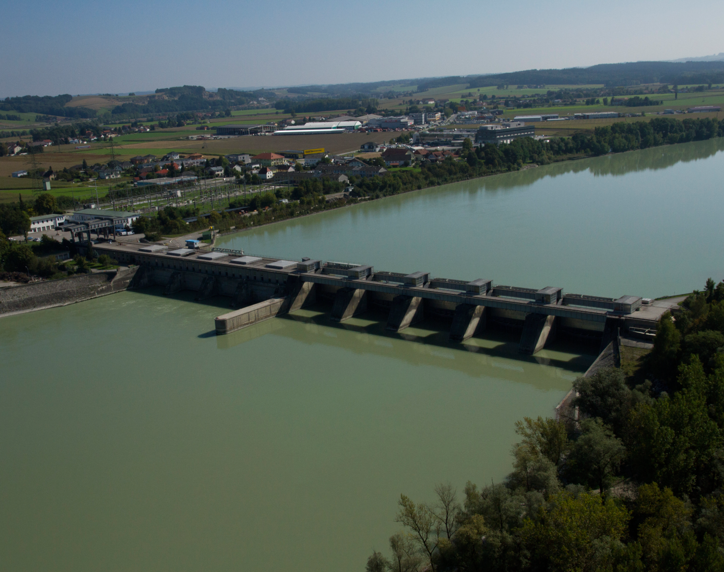 The picture shows an aerial view of the Schärding-Neuhaus hydropower plant. The power station and the blue-green Danube can be seen in the centre. On the right-hand side of the picture you can see forest and behind the power station you can see the town of Schärding and the landscape with a bright blue sky.