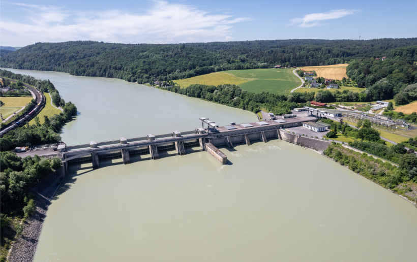 The picture shows an aerial view of the Passau-Inglings power station from the downstream side. The Inn is blue-green and therefore stands out strongly against the light and dark green meadows and forests to the left and right of the river. The course of the Inn can still be seen in the background, with forest and blue sky behind it.