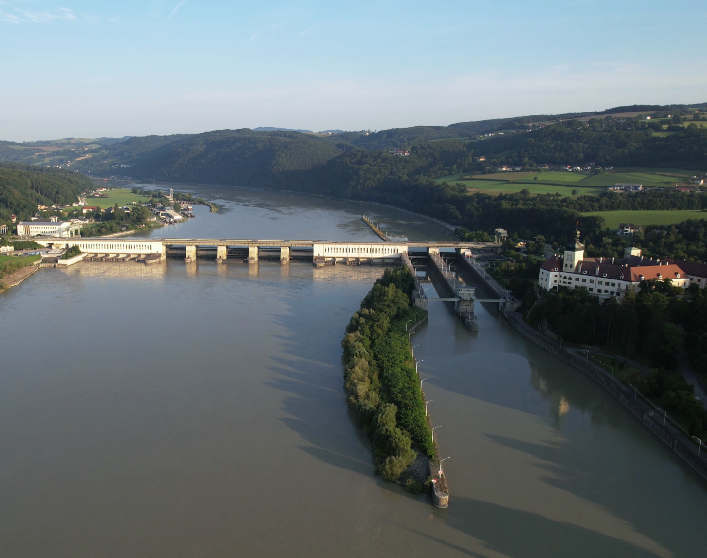 Auf dem Bild ist das Donaukraftwerk Ybbs-Persenbeug aus der Luft von der Flussabwärtsseite sichtbar. Während man im Hintergrund die schöne Landschaft mit vielen Wäldern und dem Verlauf der dunkelblauen Donau sieht, erstrahlt rechts der Ort Persenbeug im Sonnenschein. Im Hintergrund sieht man noch den blauen Himmel oberhalb der Landschaft.